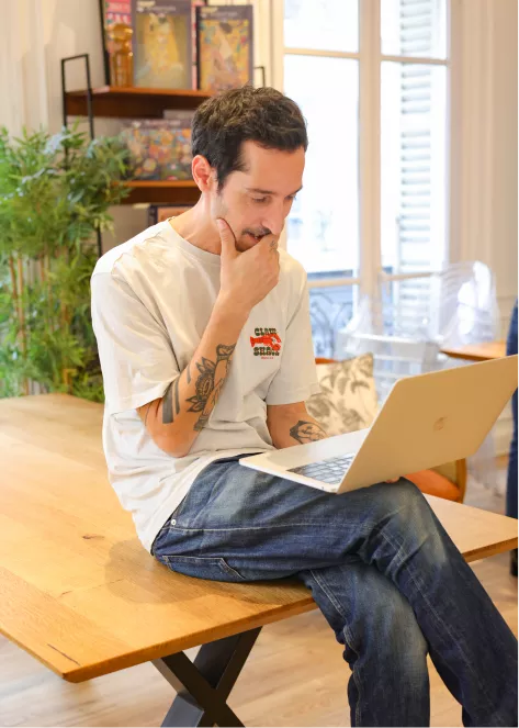 Photo de Guillaume, directeur de création, assis sur une table en train de réfléchir l'ordinateur sur les genoux, le menton dans la main