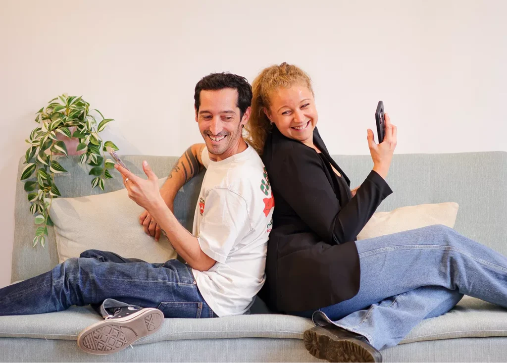 Photo de Guillaume et Pauline, respectivement directeur de création et fondatrice de l'agence, assis dos à dos sur un canapé, portable à la main, ils se jettent un regard en arrière à l'autre
