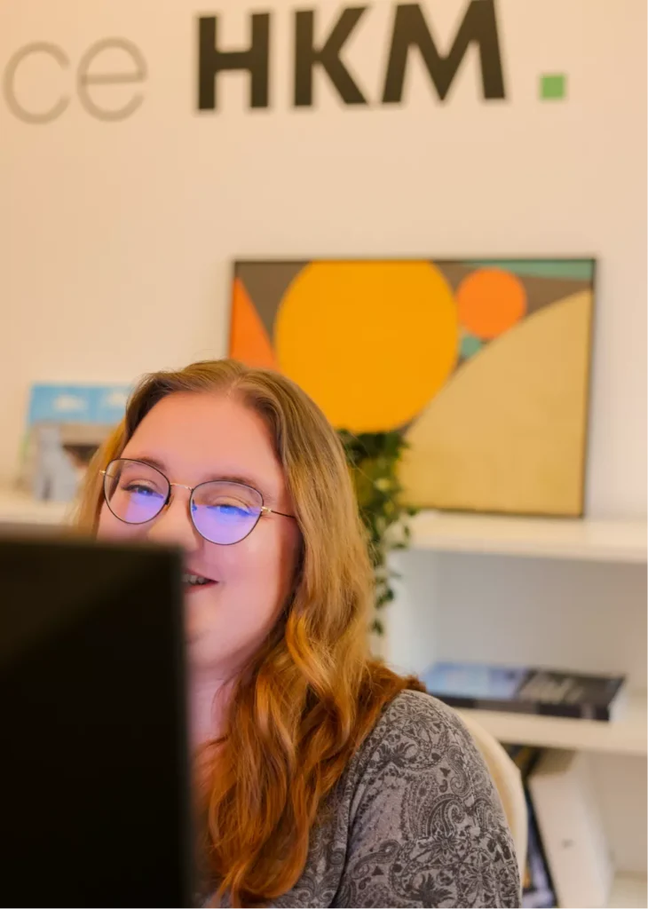 Photo de Julie, chef de projet multimédia, à son poste de travail, avec le logo HKM en arrière plan