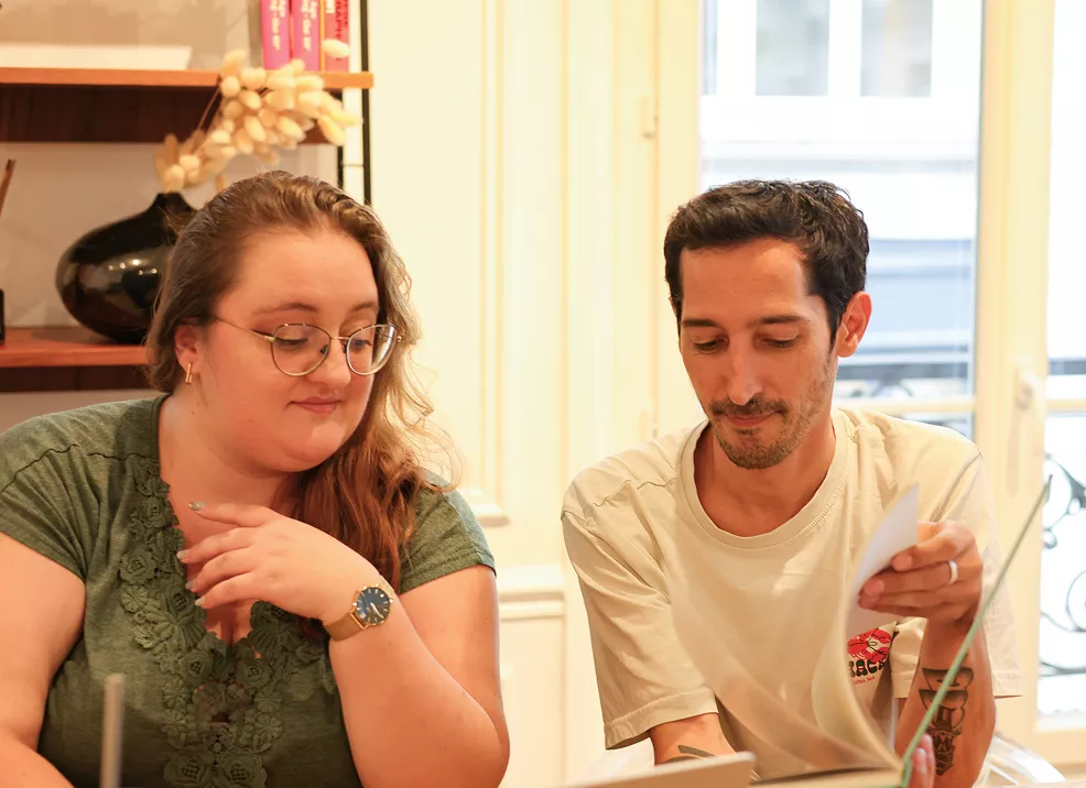 Photo de Julie, chef de projet Multimédia, et Guillaume, Directeur de création, en train de regarder le contenu d'un livre à table